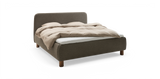Polstrad säng i textil med rundad sänggavel och träben, bäddad med beige täcke och vita lakan på frilagd bakgrund._ps