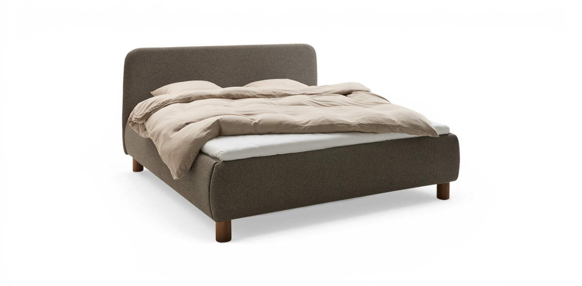Polstrad säng i textil med rundad sänggavel och träben, bäddad med beige täcke och vita lakan på frilagd bakgrund.