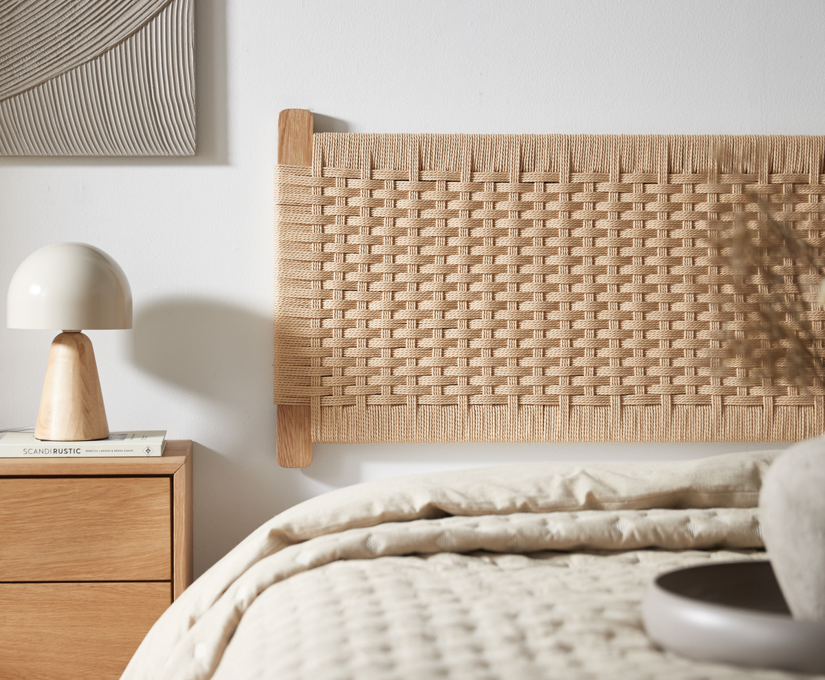 Närbild av ett ljust sovrum med flätad sänggavel i naturmaterial, trä-nattduksbord med ljus bordslampa och böcker, varav en med titeln ”SCANDI RUSTIC”; neutrala, varma toner.