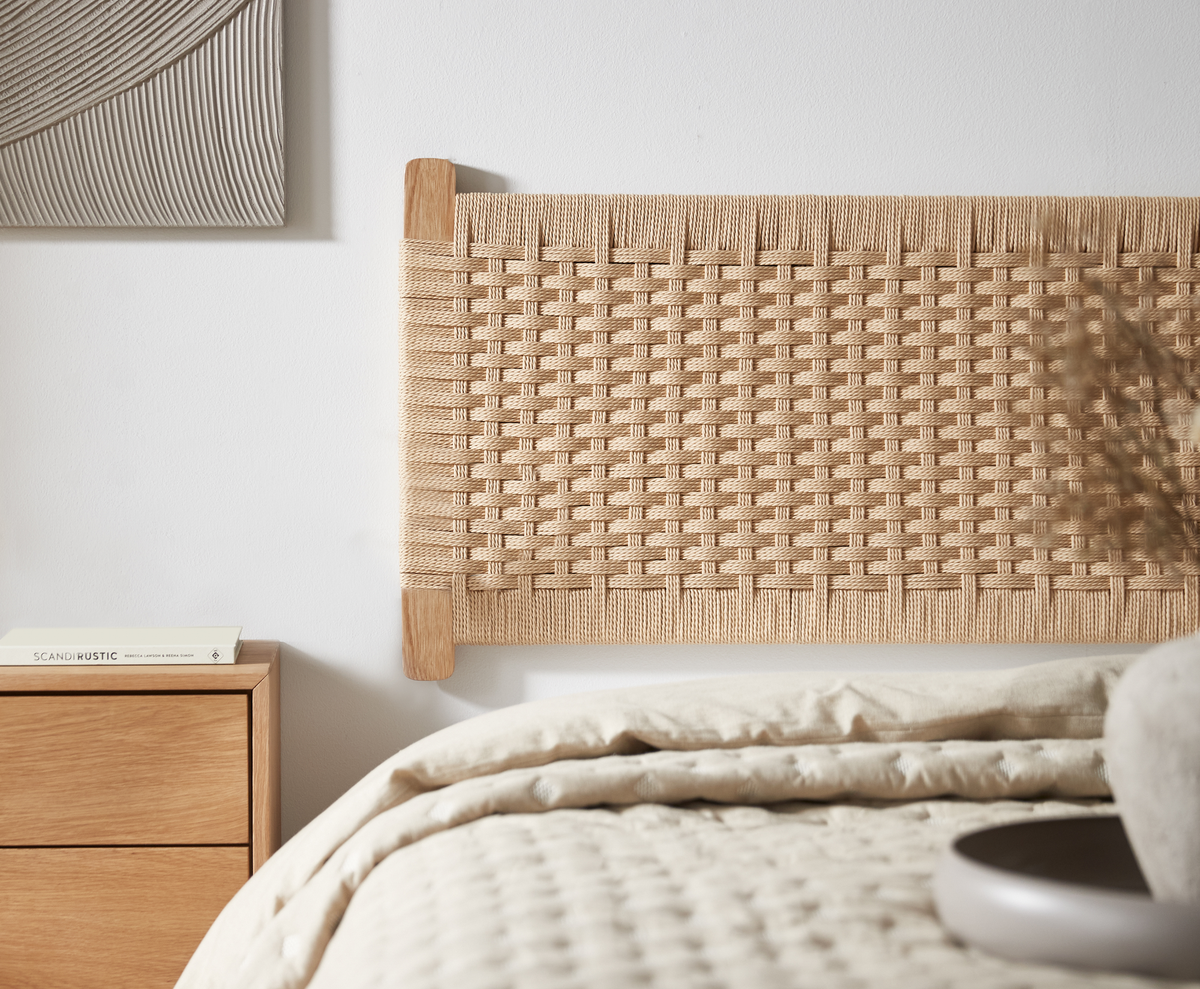 Närbild på ett sovrum med vävd naturfärgad sänggavel i trä, beige bäddning och ett nattduksbord i ljust trä med en bok med titeln SCANDI RUSTIC; ljus och luftig skandinavisk känsla.