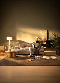 Förälder sitter i beige soffa med bebis i ett varmt upplyst vardagsrum; babytillbehör och kläder ligger utspridda på en brun matta, med golvlampa, vas med blommor, tända ljus och stor grön växt i bakgrunden.