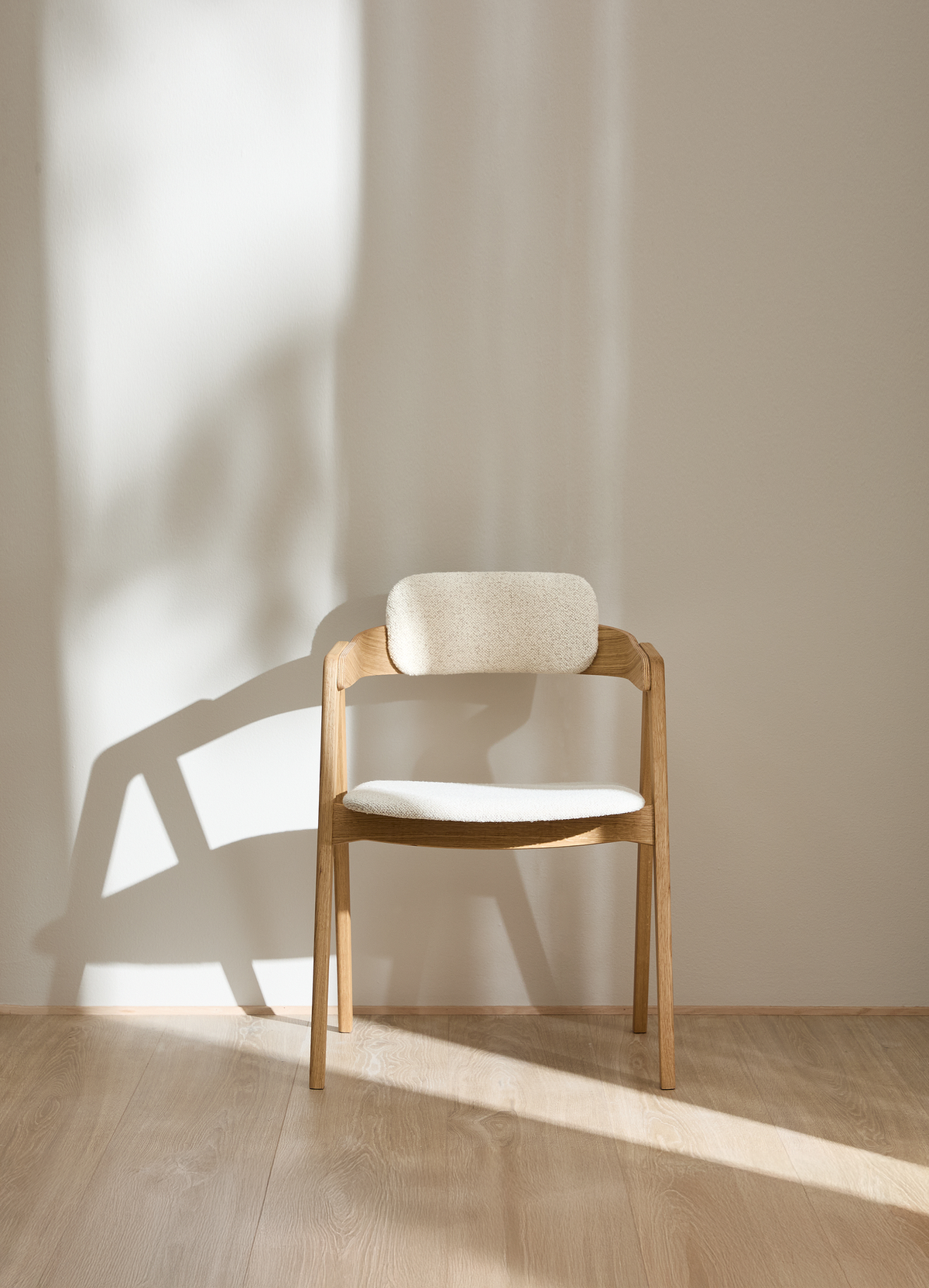Ljus trästol med ljus stoppad sits och ryggstöd i mjukt solljus mot beige vägg.