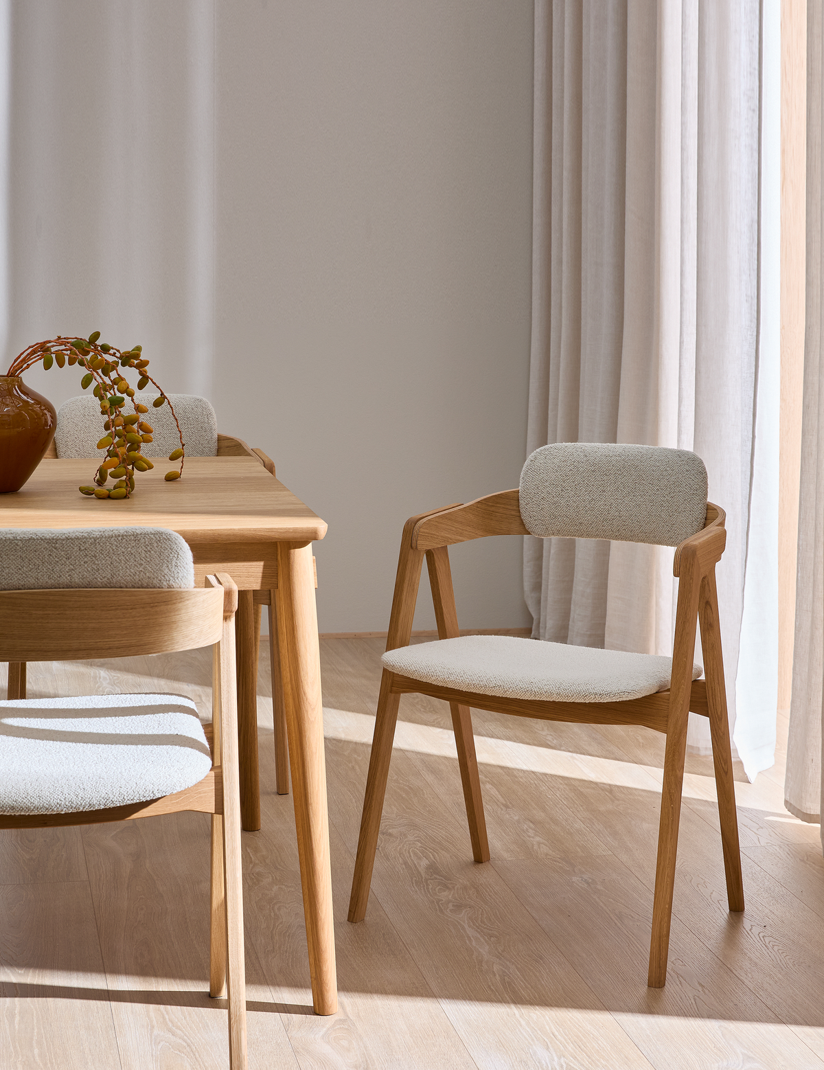 Ljust matrum med ekbord och beige stoppade stolar, solstrimmor genom ljusa gardiner och en vas med hängande grön kvist.