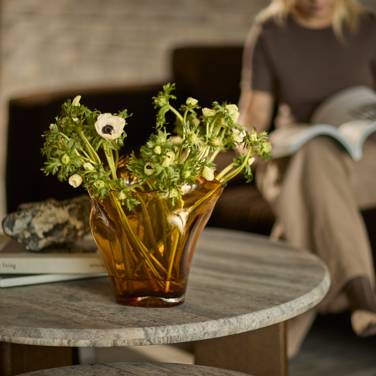 Bärnstensfärgad glasvas med vita blommor på rund stenskiva, böcker och dekorativ sten intill, suddig person som läser i soffan i bakgrunden.