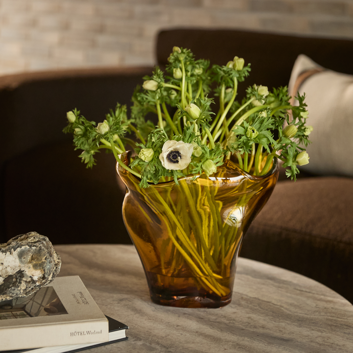 Bärnstensfärgad glasvas med vita anemoner på ett runt stenbord bredvid en sten och böcker, varav en med titeln ”HOTEL Weekend”, i en mysig vardagsrumsmiljö.
