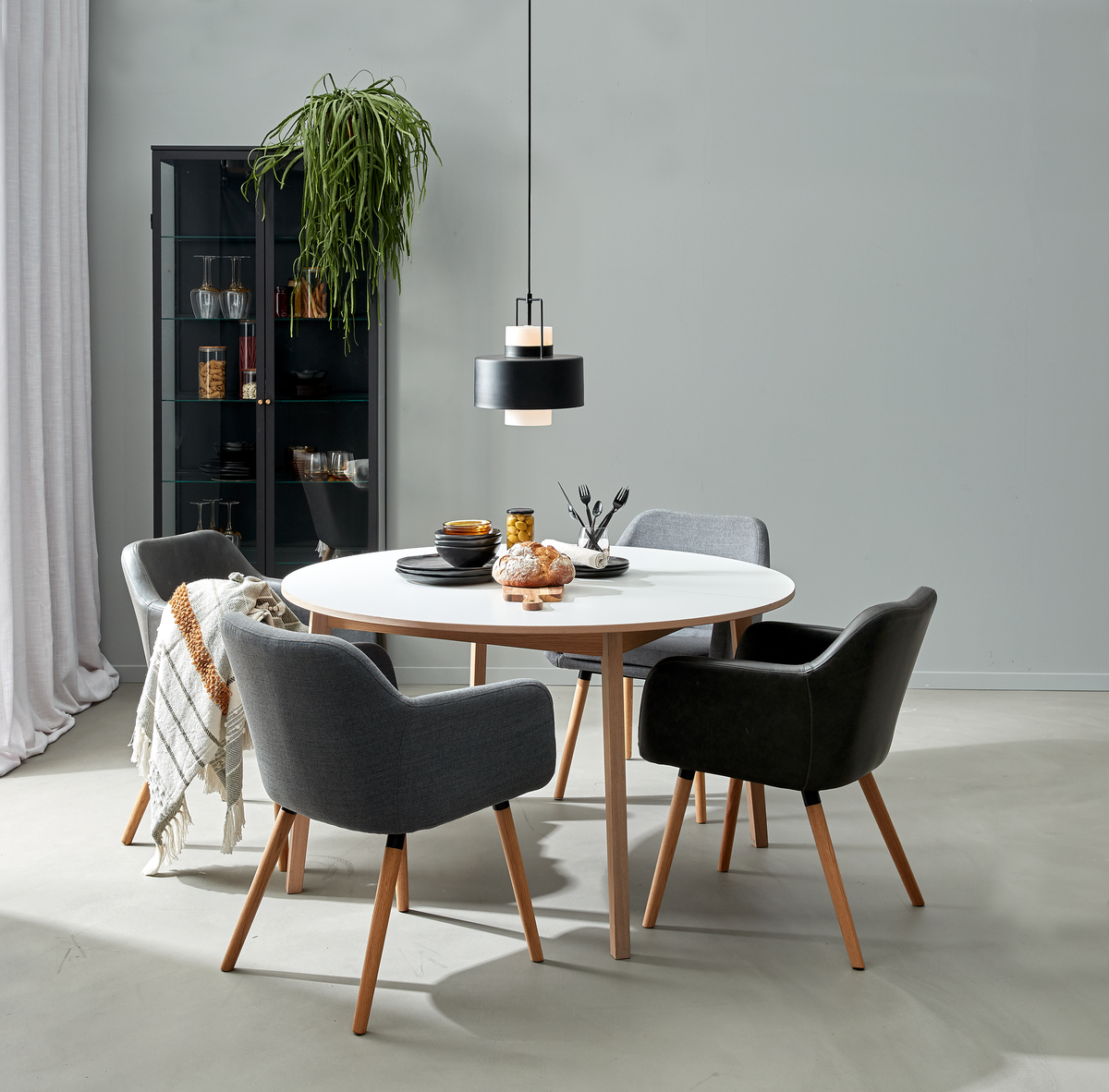 Runt vitt matbord med träben och fyra stoppade stolar under en svart pendellampa; vitrinskåp, hängande växt och filt i en minimalistisk matsal.