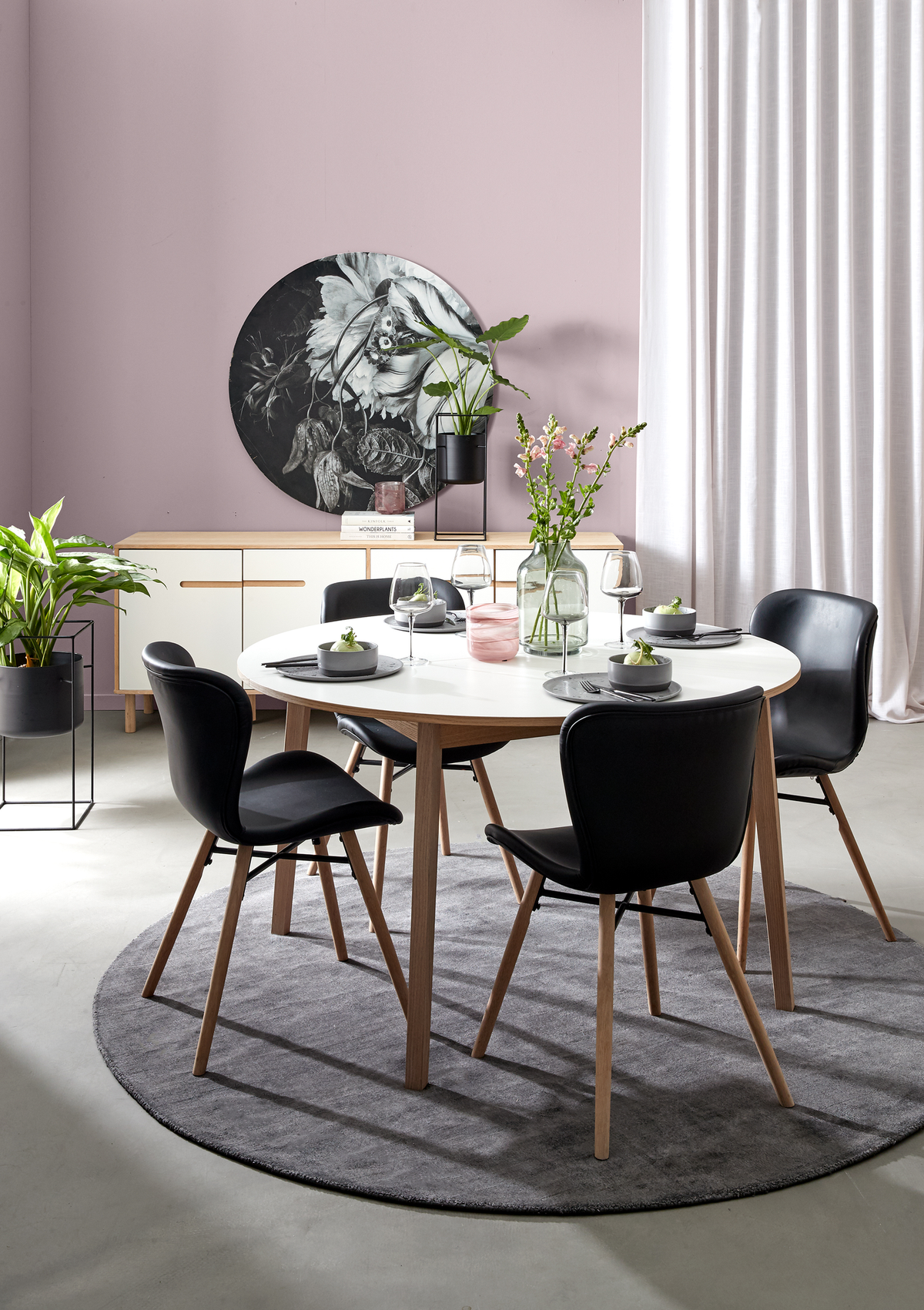 Modern matplats med runt vitt bord, svarta stolar med träben och grå rund matta; gröna växter, glasvas med blommor och dukning med vinglas framför sideboard med rund väggdekoration och ljusa gardiner i dagsljus.