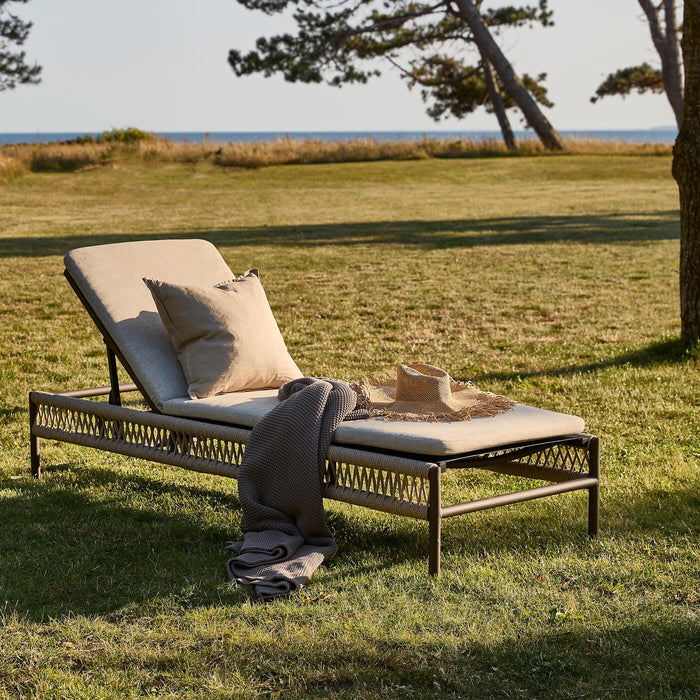 Solstol med beige dynor på gräsmatta vid havet, med kudde, filt och stråhatt i varm kvällssol.