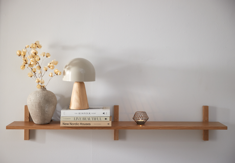 Trähylla på vit vägg med en rund betongvas med torkade kvistar, en beige bordslampa med träfot, tre böcker med titlarna The Kinfolk Home, Live Beautiful och New Nordic Houses samt en tänd liten ljuslykta i metall.