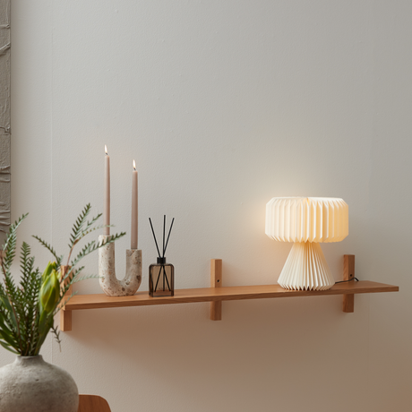 Vägmonterad trähylla med två ljus i stenljusstake, doftstickor i brun flaska och veckad bordslampa som lyser varmt; minimalistisk skandinavisk inredning.