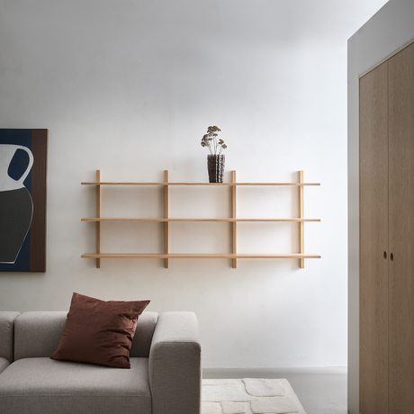 Minimalistiskt vardagsrum i ljusa toner med väggmonterad hylla i ljust trä, brun glasvas med torkade blommor, grå soffa med brun kudde, abstrakt tavla och mjuk matta.