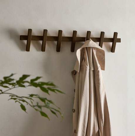 Vägghängd träkroklist med en beige trenchcoat på en ljus vägg, suddig grön växt i förgrunden.