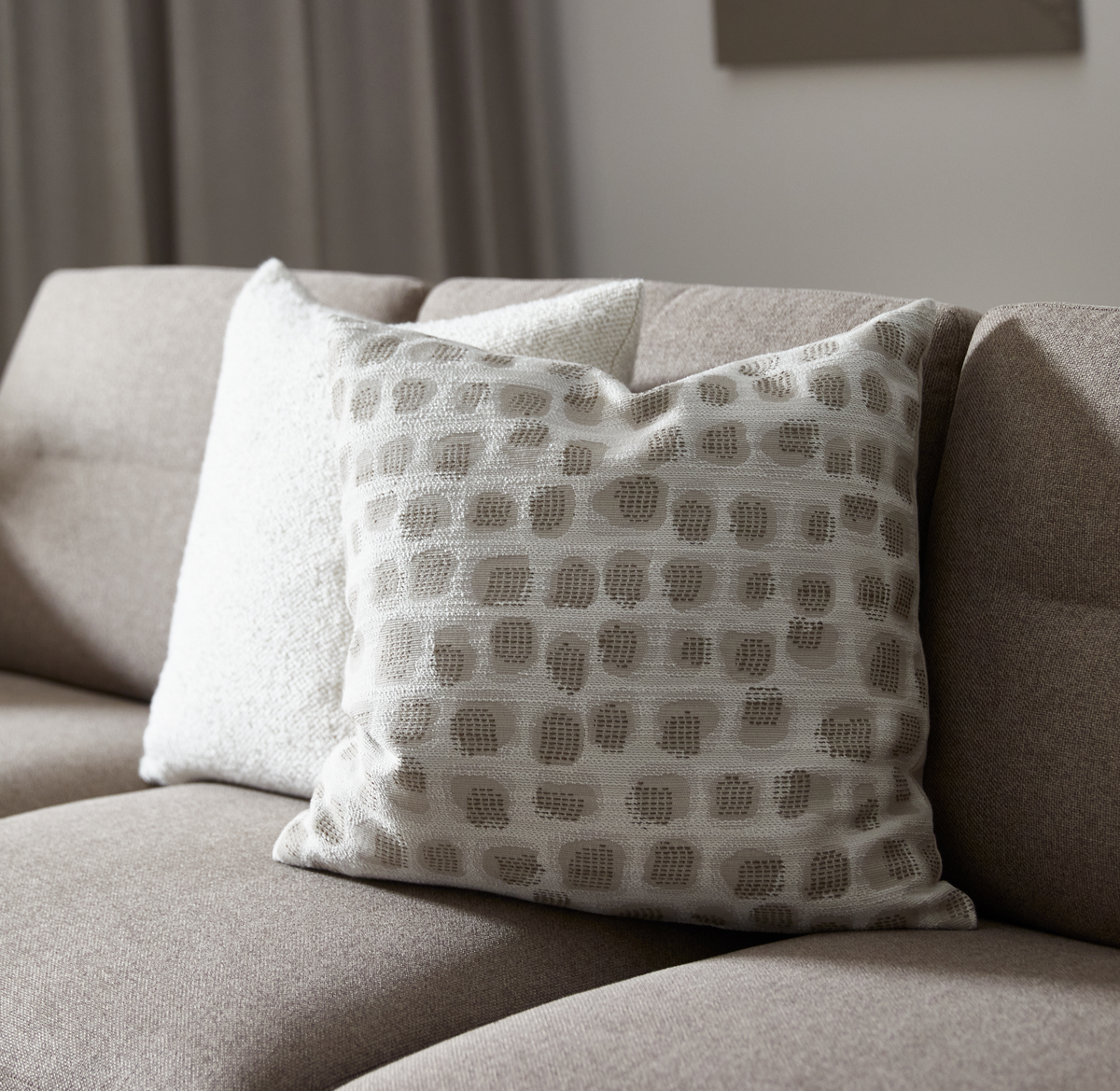 Beige soffa med två prydnadskuddar i neutrala toner; främre kudden har ett ljusbeige mönster med strukturerade prickar.