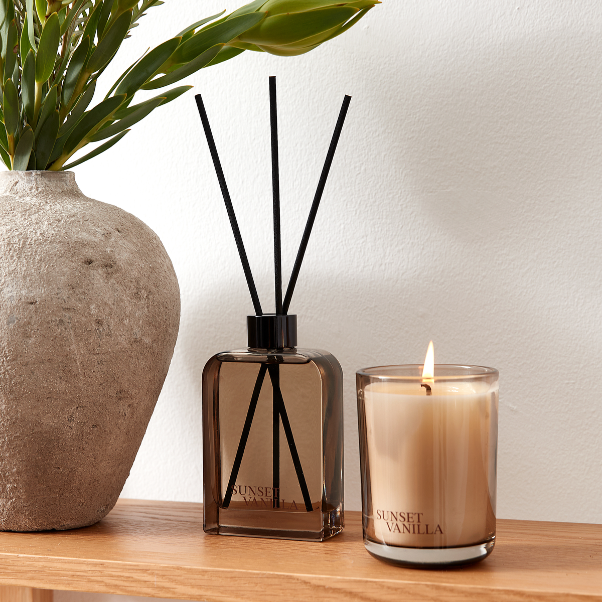 Doftspridare och tänt ljus i brunt glas med Sunset Vanilla på en trähylla bredvid en stenkänslig vas med gröna blad.