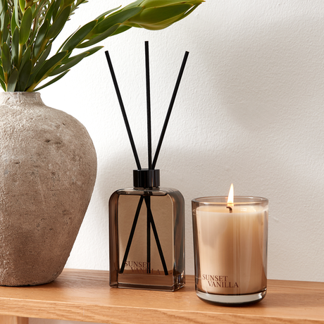 Doftspridare och tänt ljus i brunt glas med Sunset Vanilla på en trähylla bredvid en stenkänslig vas med gröna blad.
