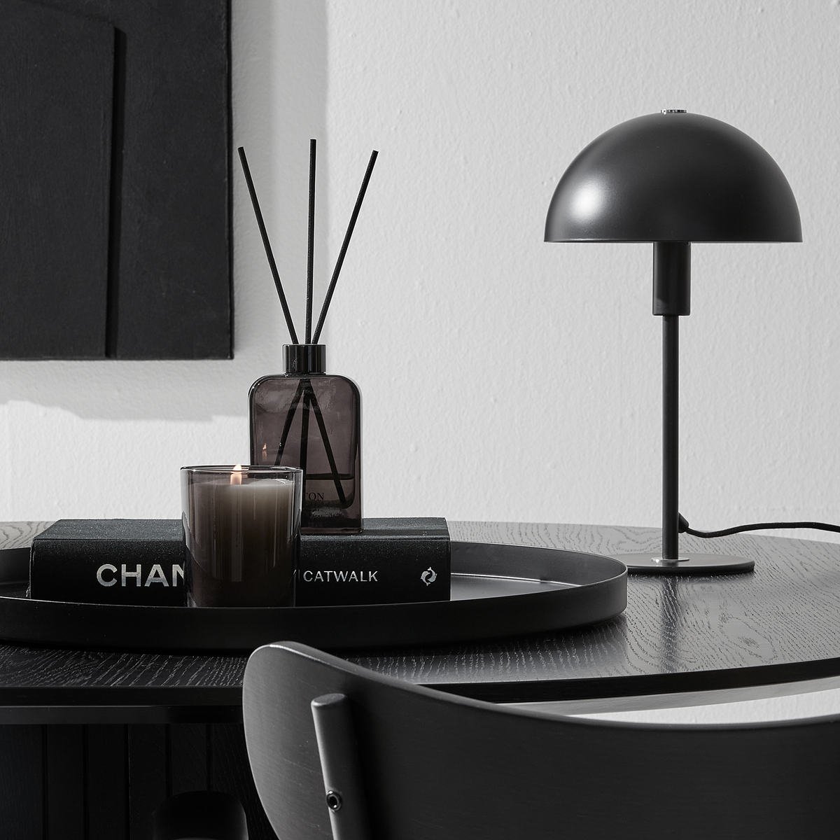 Svart bordsvy med kupolformad bordslampa, doftpinnar och tänt ljus på en bricka ovanpå böckerna CHANEL och CATWALK.