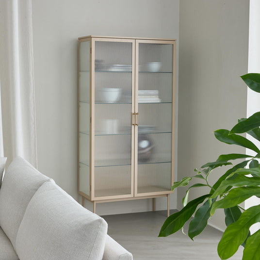 Ljust vitrinskåp med räfflade glasdörrar i ett minimalistiskt vardagsrum, med vita skålar och böcker på glas-hyllor och en grön växt bredvid.