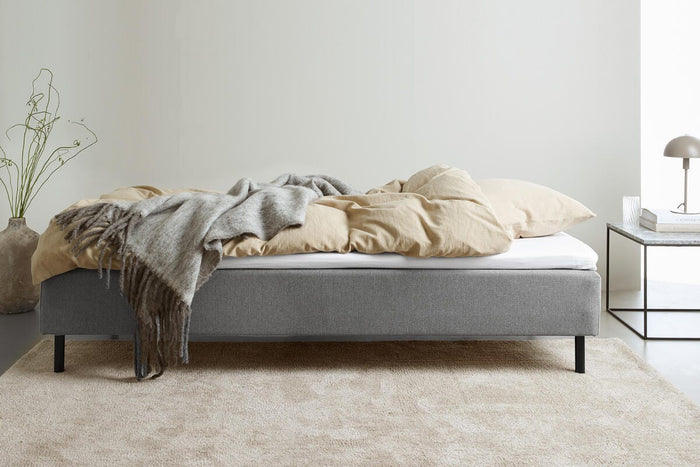 Grå stoppad säng med beige bäddset och grå pläd; nattduksbord med lampa och böcker i ett ljust, minimalistiskt sovrum.