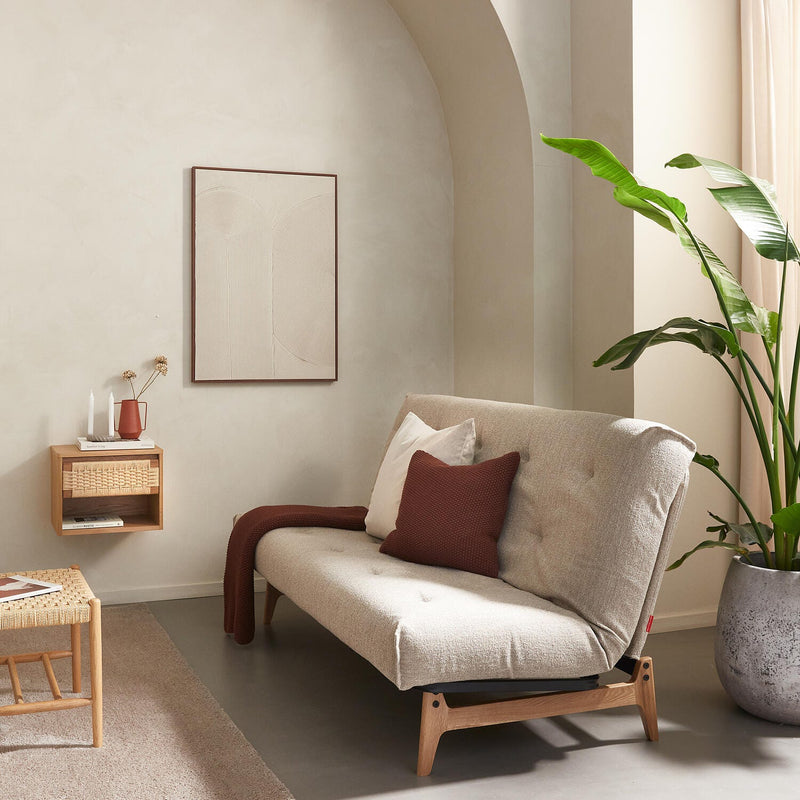 Ljus skandinavisk vardagsrumsmiljö med beige soffa på träben, roströd kudde och pläd, stor grön växt, svävande hylla med vas och ljus samt flätat soffbord i neutrala toner.