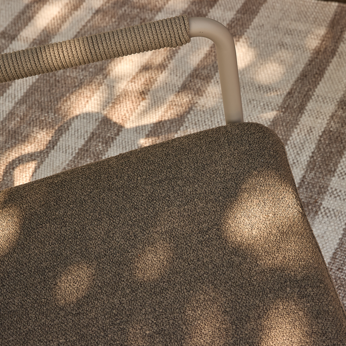 Närbild av en beige utomhusstol med repklädd metallram och texturerad sits ovanpå en randig matta i solfläckat dagsljus.