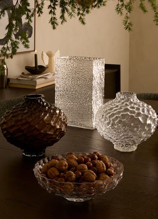 Texturerade glasvaser och skålar på ett mörkt matbord, en skål med nötter, varm belysning med grön kvist ovanför samt ljus och väggkonst i bakgrunden.