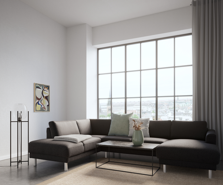 Ljust vardagsrum med mörk hörnsoffa framför stora spröjsade fönster med stadsvy, soffbord i sten med grön vas och torkade kvistar, gråa kuddar och pläd, golvlampa och abstrakt tavla.