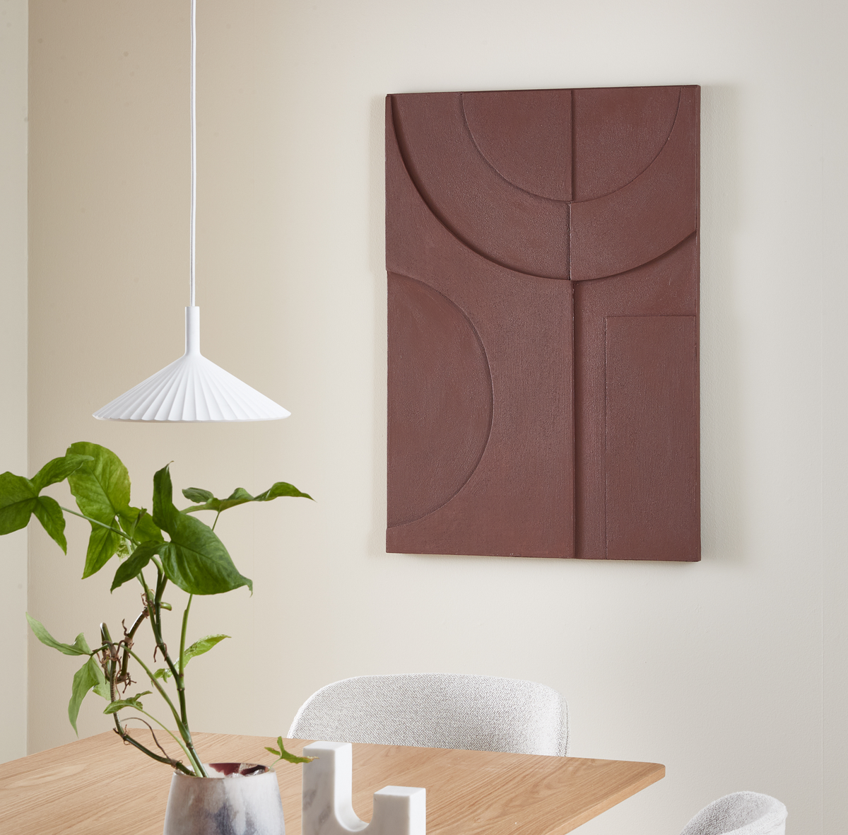 Matplats med vit plisserad pendellampa, grön växt i vas på träbord och brun geometrisk väggpanel på ljus vägg.