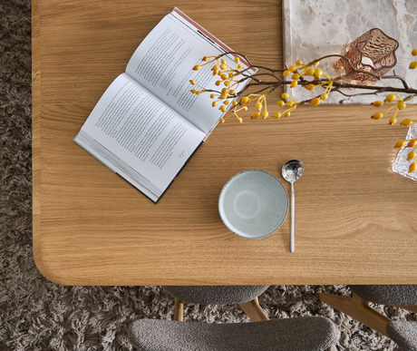 Stilrent topview över ett matbord i ek med öppen bok med text, en ljus skål med sked, en kvist med gula bär och en glaslykta på marmorfat; mjukt ryatäcke och stoppade stolar skymtar.