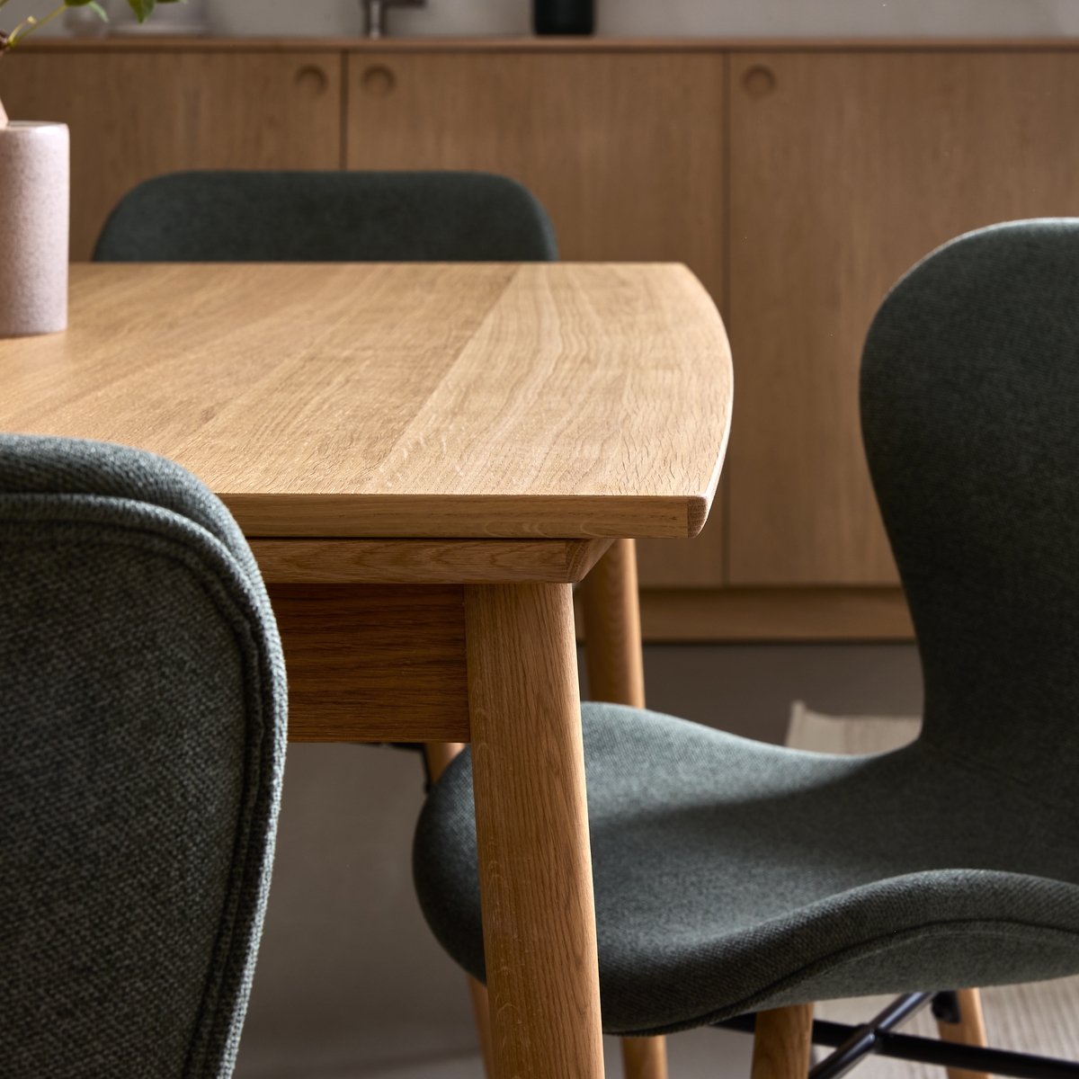 Närbild av matbord i ek med rundade kanter och gröna, klädda stolar framför ett sideboard i trä.