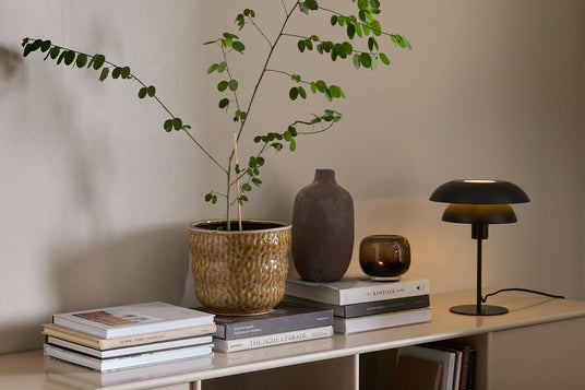 Beigefärgad sideboard med grön krukväxt, keramikvas, tänd ljuskopp och svart bordslampa ovanpå staplade böcker som The Kinfolk Home och The Home Upgrade mot en neutral vägg.
