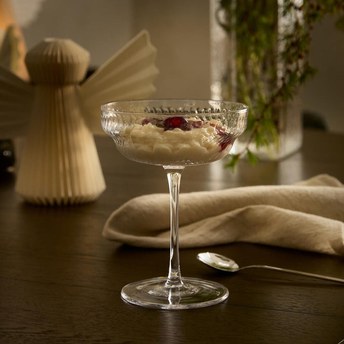 Dessert serverad i en räfflad coupe-glas på ett mörkt träbord, med linneservett och sked intill; suddig vas med gröna kvistar och en pappersängel i bakgrunden i varm belysning.