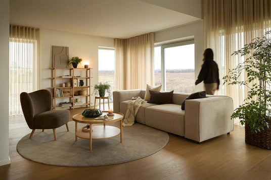 Ljus skandinavisk vardagsrum med beige hörnsoffa, brun fåtölj och rund matta; stora fönster med skira gardiner, trähyllor och träbord med grön skål och tändt ljus; en person i rörelse vid fönstret.