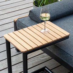 Litet sidobord i trä med svart metallram bredvid en grå utomhussoffa på trädäck; ett glas vitt vin på bordet och en öppen bok samt kuddar på soffan.