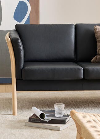 Svart lädersoffa med ljus trästomme och en beige strukturkudde; framför ligger en hög coffeetable-böcker – bl.a. ”Axel Vervoordt: Portraits of Interiors” – med ett räfflat glas ovanpå; vävd loungestol och neutral matta i ett ljust vardagsrum.