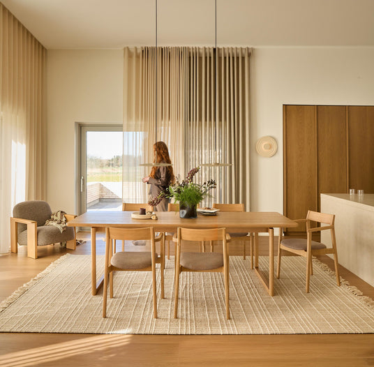 Ljust skandinaviskt matrum med träbord och fyra stolar på beige matta, skira gardiner, pendellampor och vas med blommor; person vid altandörr.
