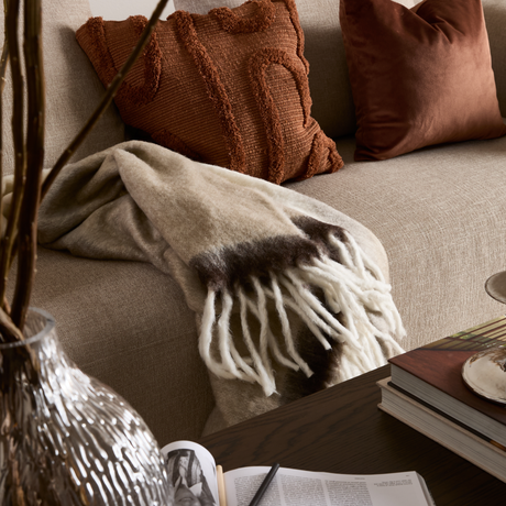 Beige soffa med rostbruna kuddar, ullpläd med fransar i beige och brunt, glasvas med kvistar och böcker på ett mörkt soffbord – mysig vardagsrumsdetalj.