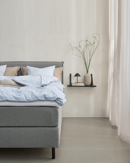 Minimalistiskt sovrum med grå säng, ljusblått bäddset och beige kuddar; bredvid sitter en svart väggmonterad hylla med liten lampa, böcker och en vas med långsmala kvistar vid ljusa gardiner.