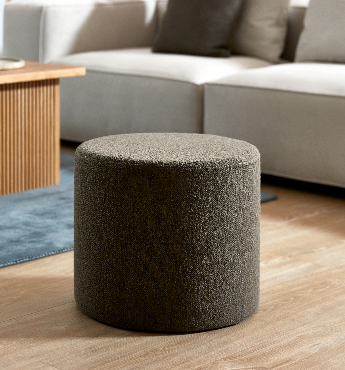 Rund puff i mörkbeige, texturerat tyg i ett vardagsrum med ljus soffa, träbord och matta.