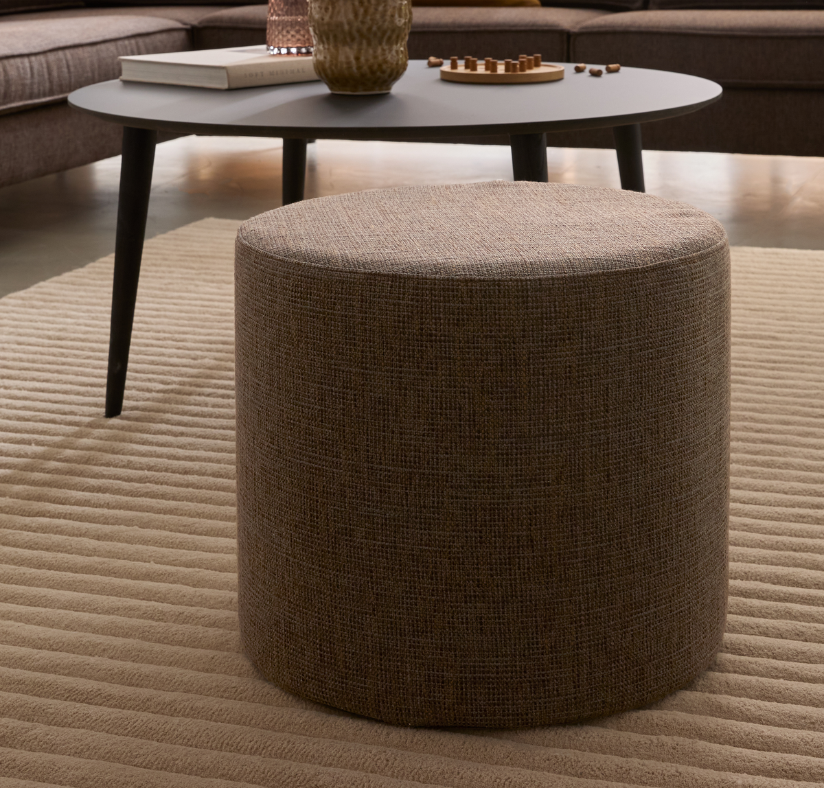 Beige cylinderformad sittpuff på en ribbad beige matta framför ett mörkgrått soffbord med bok, vas och liten spelbricka.