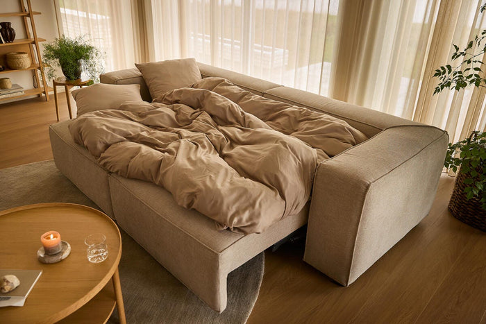 Beige bäddsoffa uppfälld med sandfärgat påslakan i ett ljust vardagsrum med skira gardiner, runt träbord med tändt ljus och gröna växter.