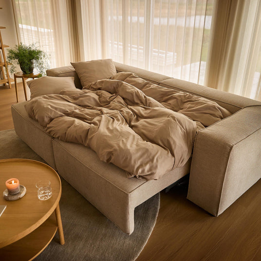 Beige bäddsoffa uppfälld med sandfärgat påslakan i ett ljust vardagsrum med skira gardiner, runt träbord med tändt ljus och gröna växter.