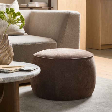 Brun rund sittpuff framför beige soffa, marmorbord med vågig vas och gröna kvistar i ett ljust vardagsrum.