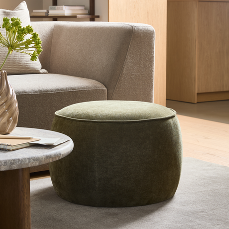 Grön rund sittpuff framför beige soffa, marmorbord med vas och böcker i ljust vardagsrum.