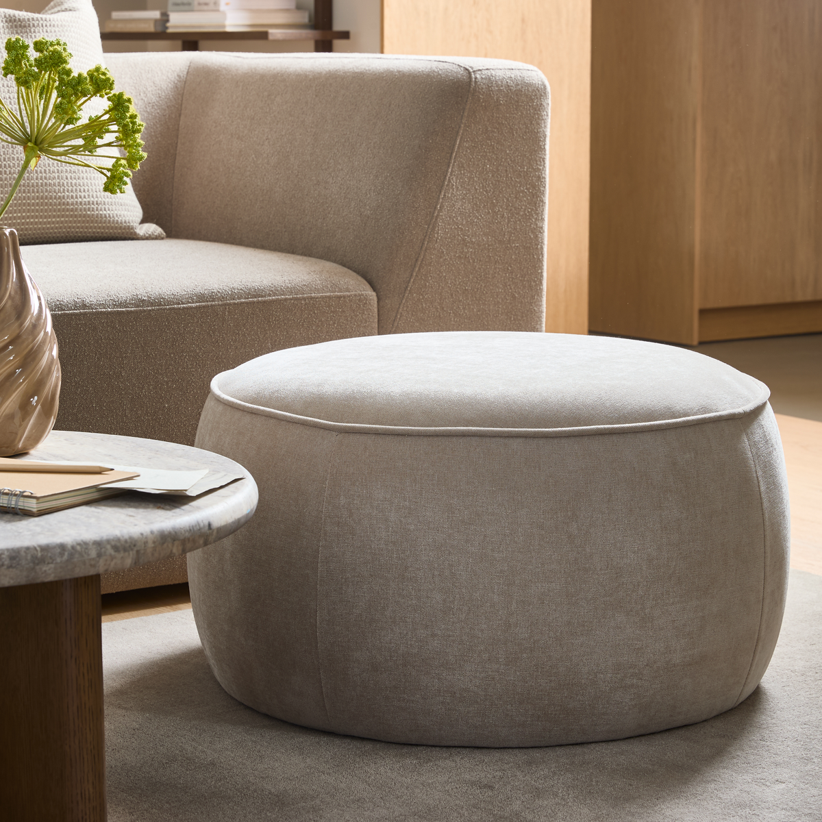 Rund beige tygpuff i ett modernt skandinaviskt vardagsrum bredvid en kurvad soffa och ett stenlikt soffbord.