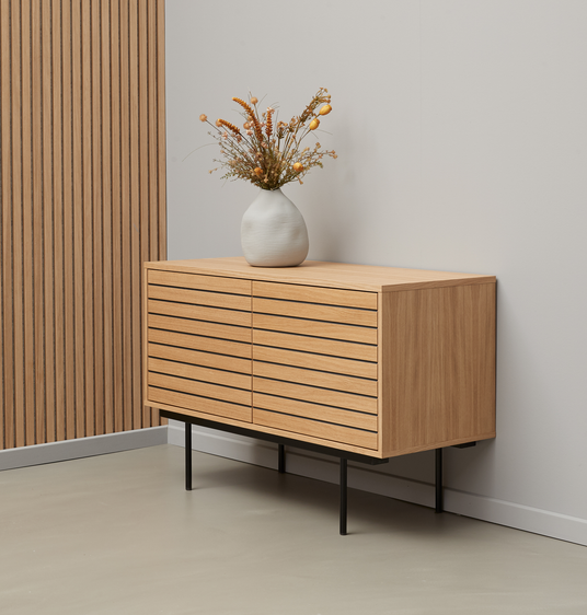 Låg sideboard i ljus ek med horisontella ribbfronter på svarta ben, dekorerad med vit vas med torkade blommor mot ljus vägg.