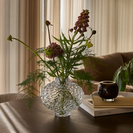 Glasvas med struktur och blandade blommor på ett träbord bredvid en brun ljuslykta på två böcker, där den nedersta har titeln ”barefoot living”, i mjukt dagsljus med gardiner i bakgrunden.
