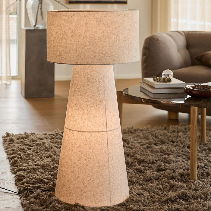 Beige golvlampa med koniskt, tygklätt stativ och stor cylindrisk lampskärm i ett skandinaviskt vardagsrum med ryamatta och soffbord.