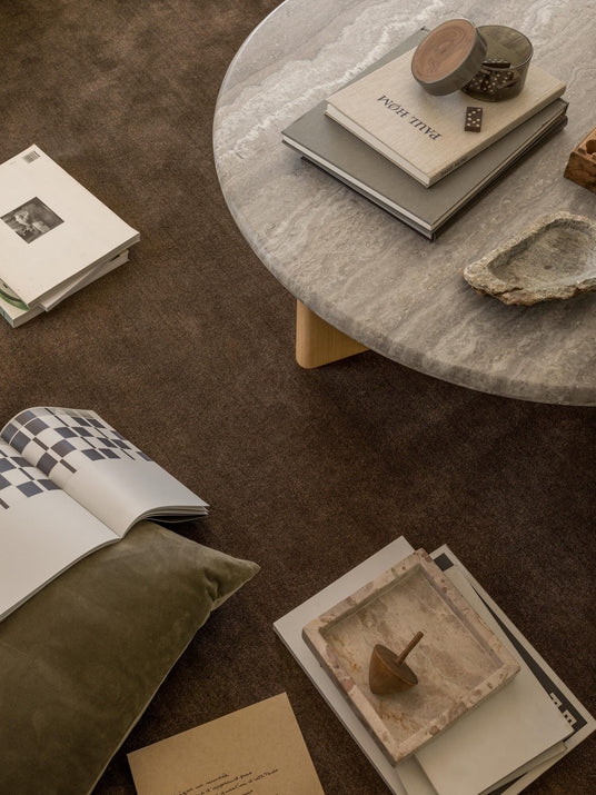 Rundt soffbord i sten på brun matta med staplade böcker (en med titeln ”PAUL HOME”), öppet magasin med schackrutigt mönster, liten behållare med dominobrickor och en bricka i sten med en träsnurra; grön sammetskudde intill.