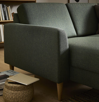 Närbild av grön tygsoffa med träben i vardagsrum; korgpall med anteckningsbok och boken A Modern Way to Live bredvid, bokhylla i bakgrunden.
