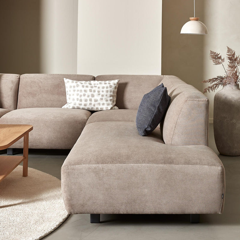 Beige hörnsoffa i tyg med två prydnadskuddar, runt ljust ryamatta, träsoffbord, pendellampa och stor vas med torkade grenar i ett ljust vardagsrum.